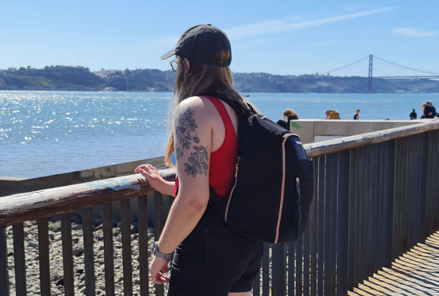 A woman is wearing the travel hack backpack, standing in front of the river in Lisbon with a bridge in the background